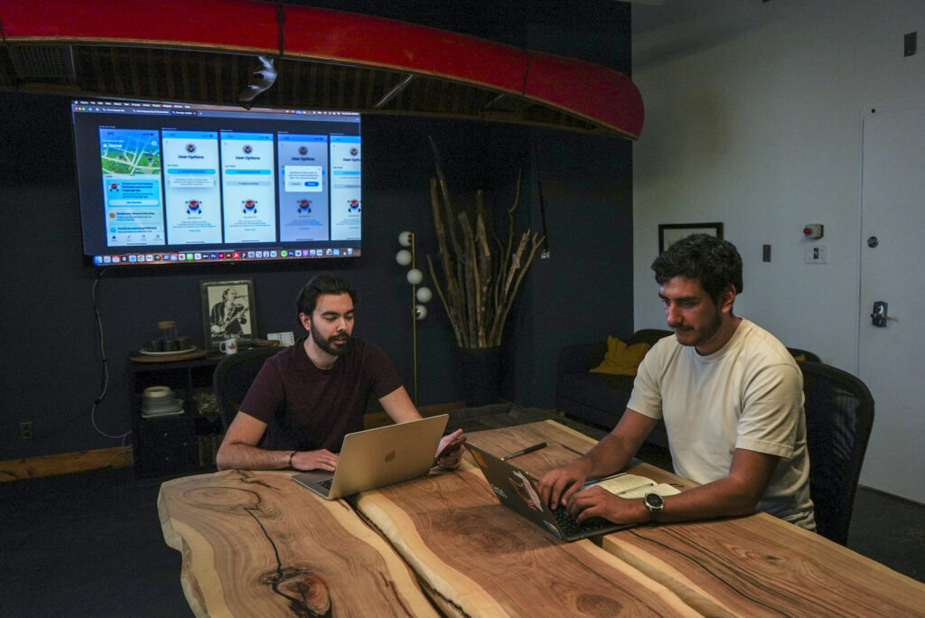 two men sitting at a table chatting with their laptops open.